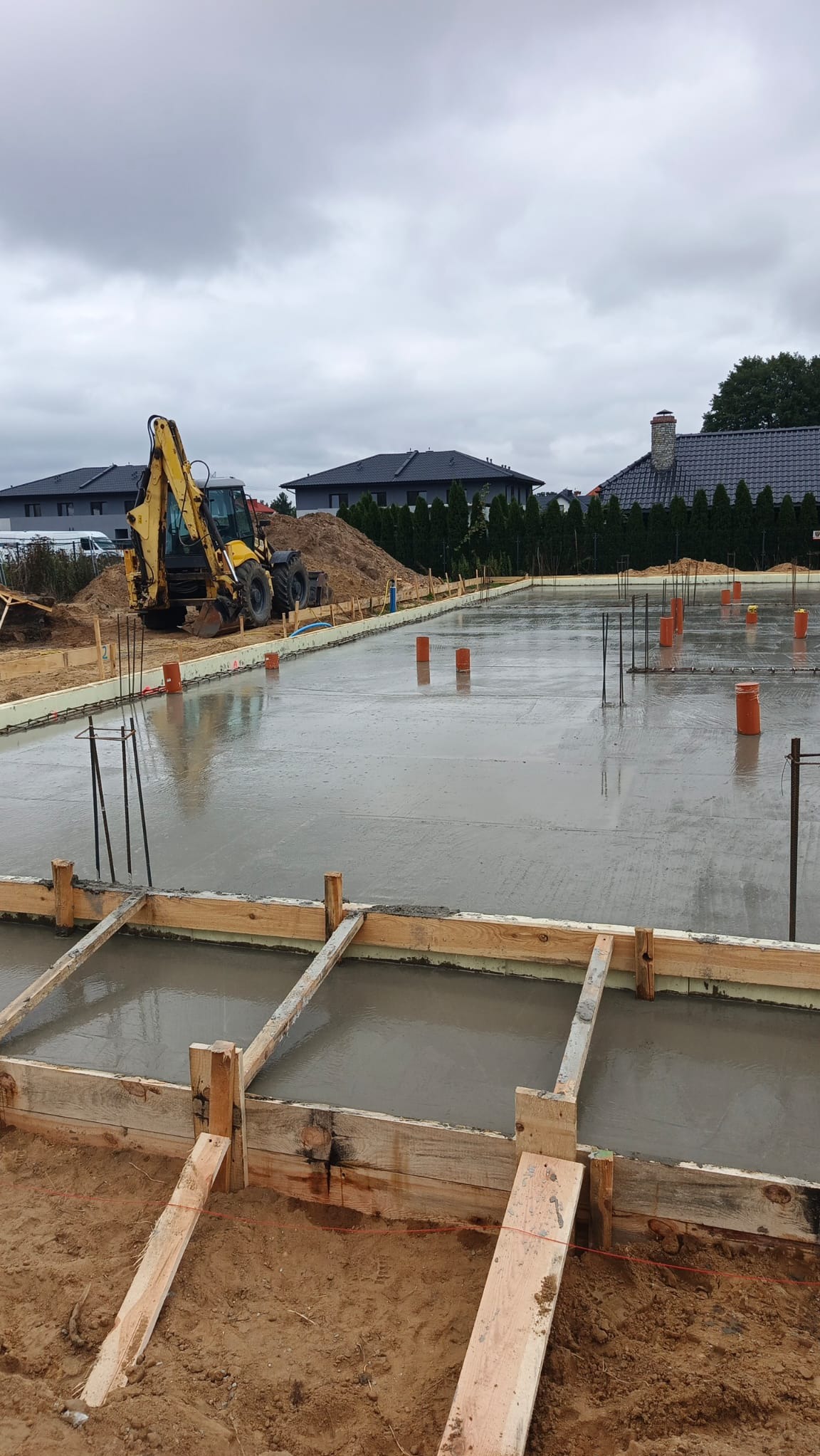 Zalany fundament pod budowę domu jednorodzinnego. Widoczne szalunki, wystające pręty zbrojeniowe i rury. W tle koparka i hałda ziemi.