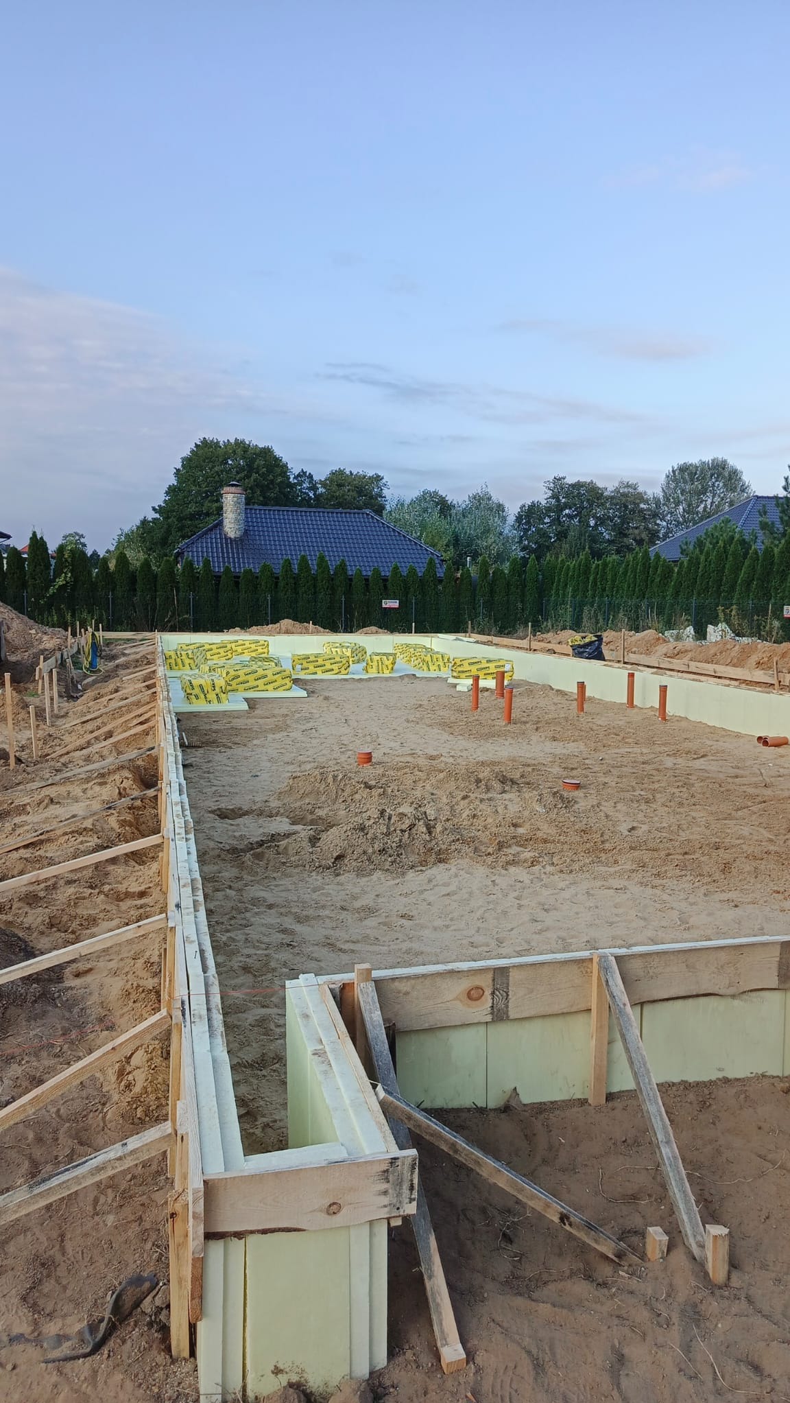 Fundamenty domu w trakcie budowy: szalunek z desek, izolacja termiczna, piasek. Widoczne wystające rury i zapakowane materiały izolacyjne. Tło: dom i drzewa.