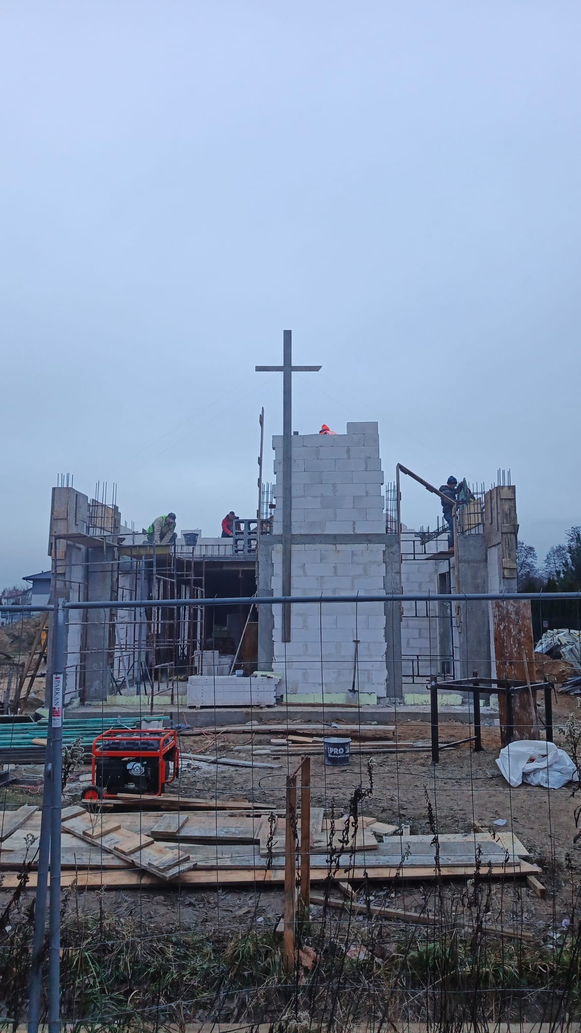 Budowa kościoła w toku: murarze pracują na rusztowaniach, wznosząc ściany z białych bloczków. W tle widoczny krzyż i pochmurne niebo.