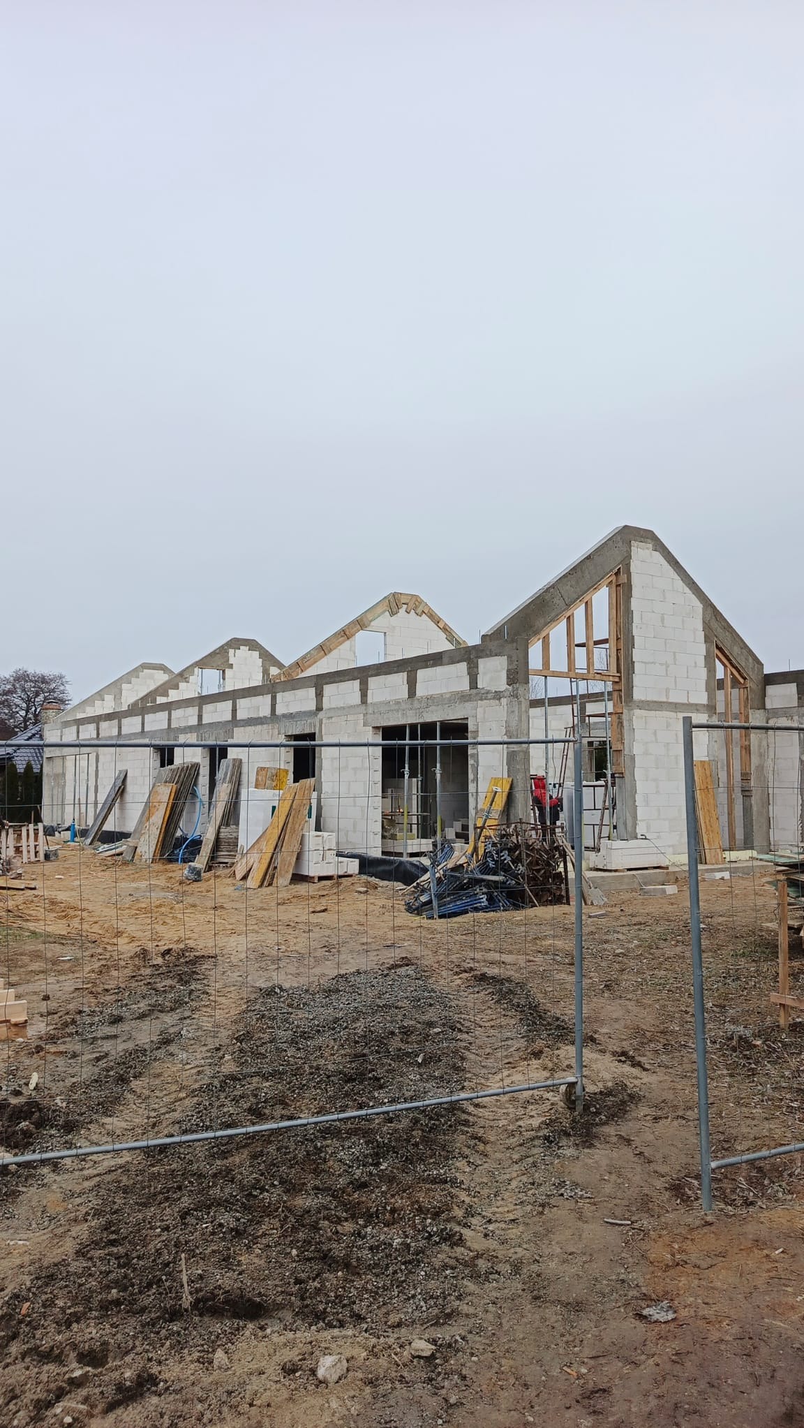 Budowa domu z betonu komórkowego w stanie surowym otwartym, widoczne ściany, nadproża, drewniane szalunki okienne, teren budowy zabezpieczony siatką.