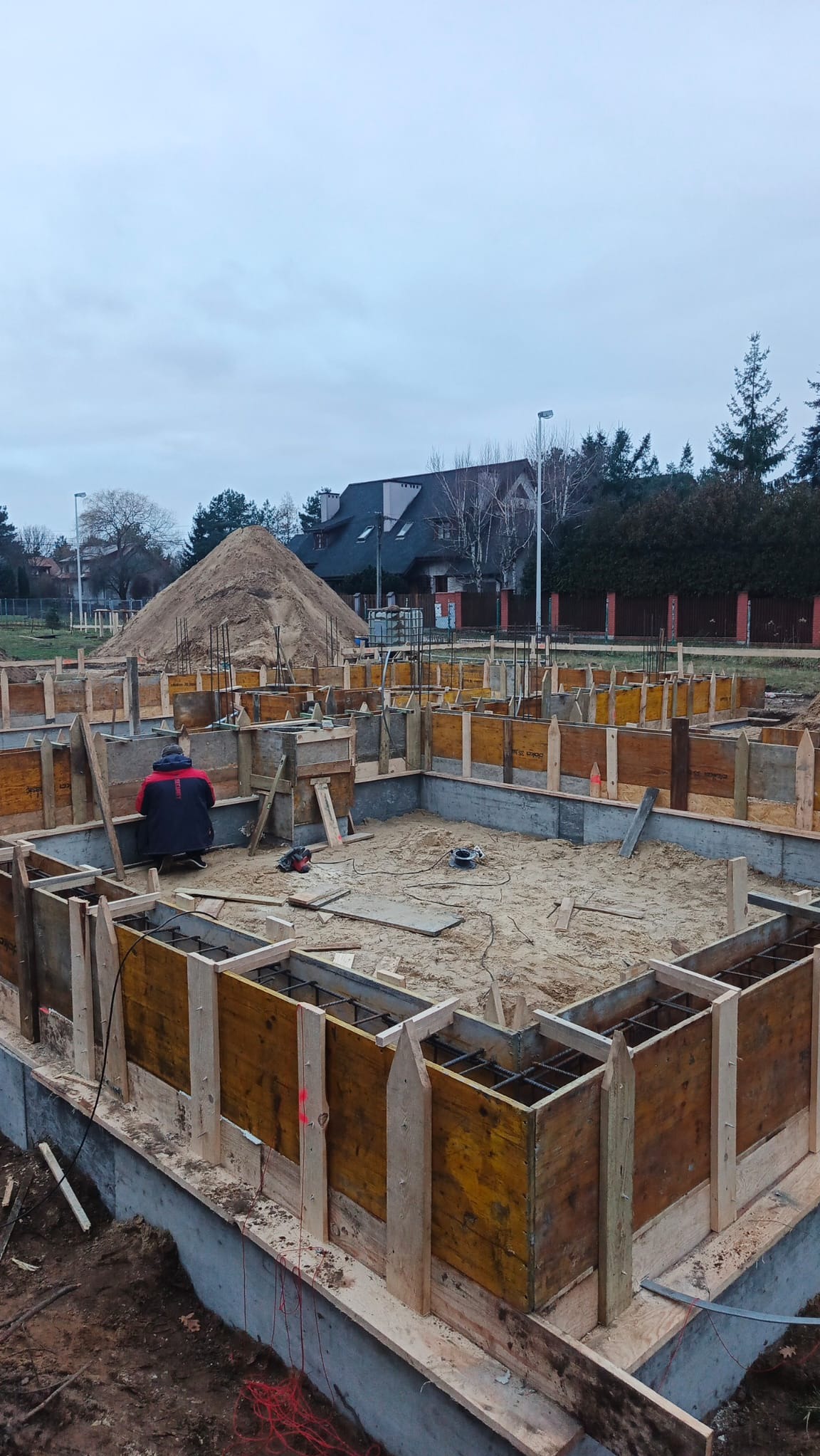 Zbrojenie i szalunek fundamentów domu jednorodzinnego. Widoczny pracownik w trakcie pracy, narzędzia oraz hałda piasku w tle. Ujęcie z góry, pochmurny dzień.