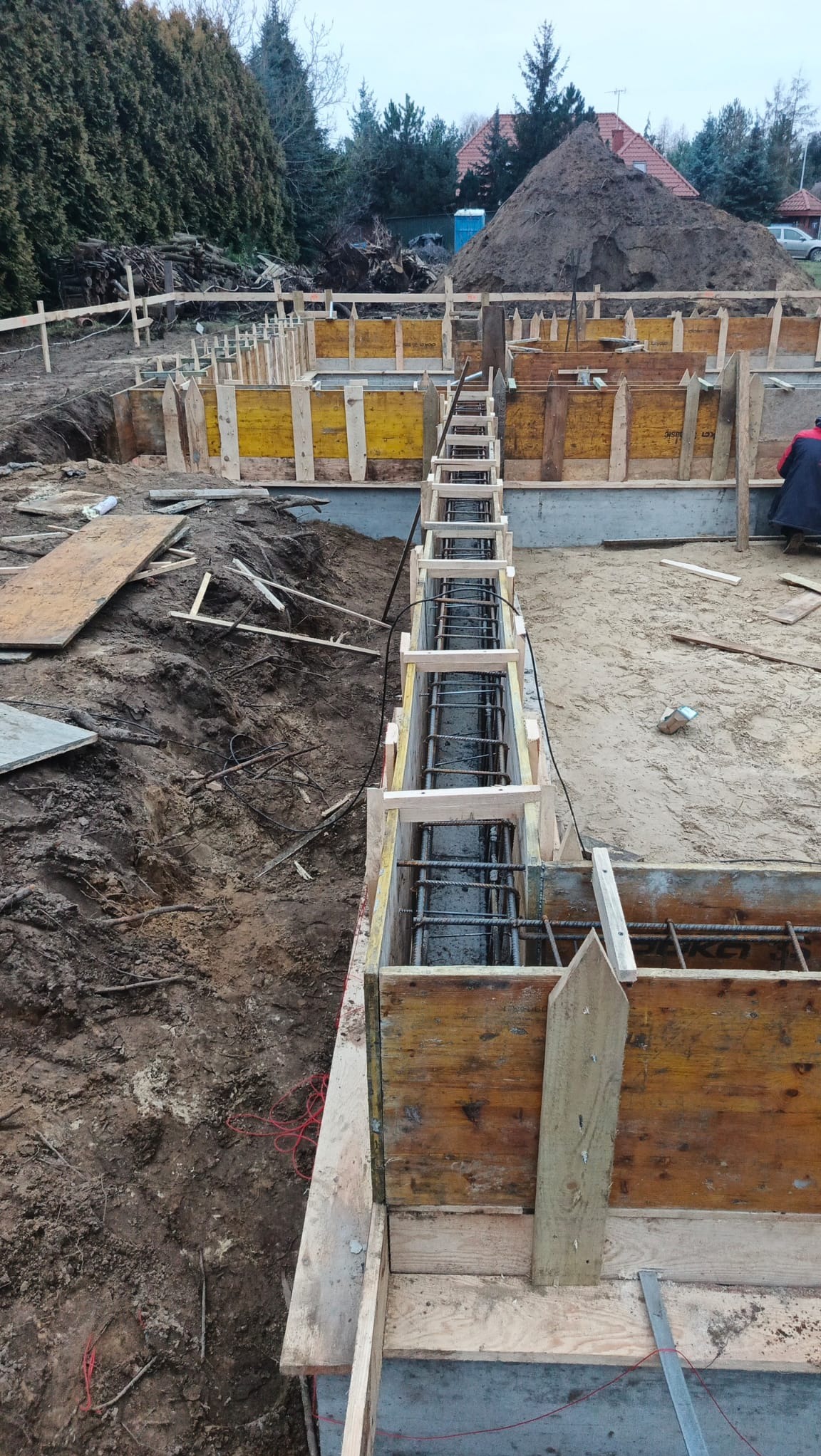 Zbrojenie fundamentów w drewnianym szalunku, widoczne pręty i beton. W tle ziemia, drzewa i fragment budynku. Ujęcie z góry, ukazujące detale konstrukcji.