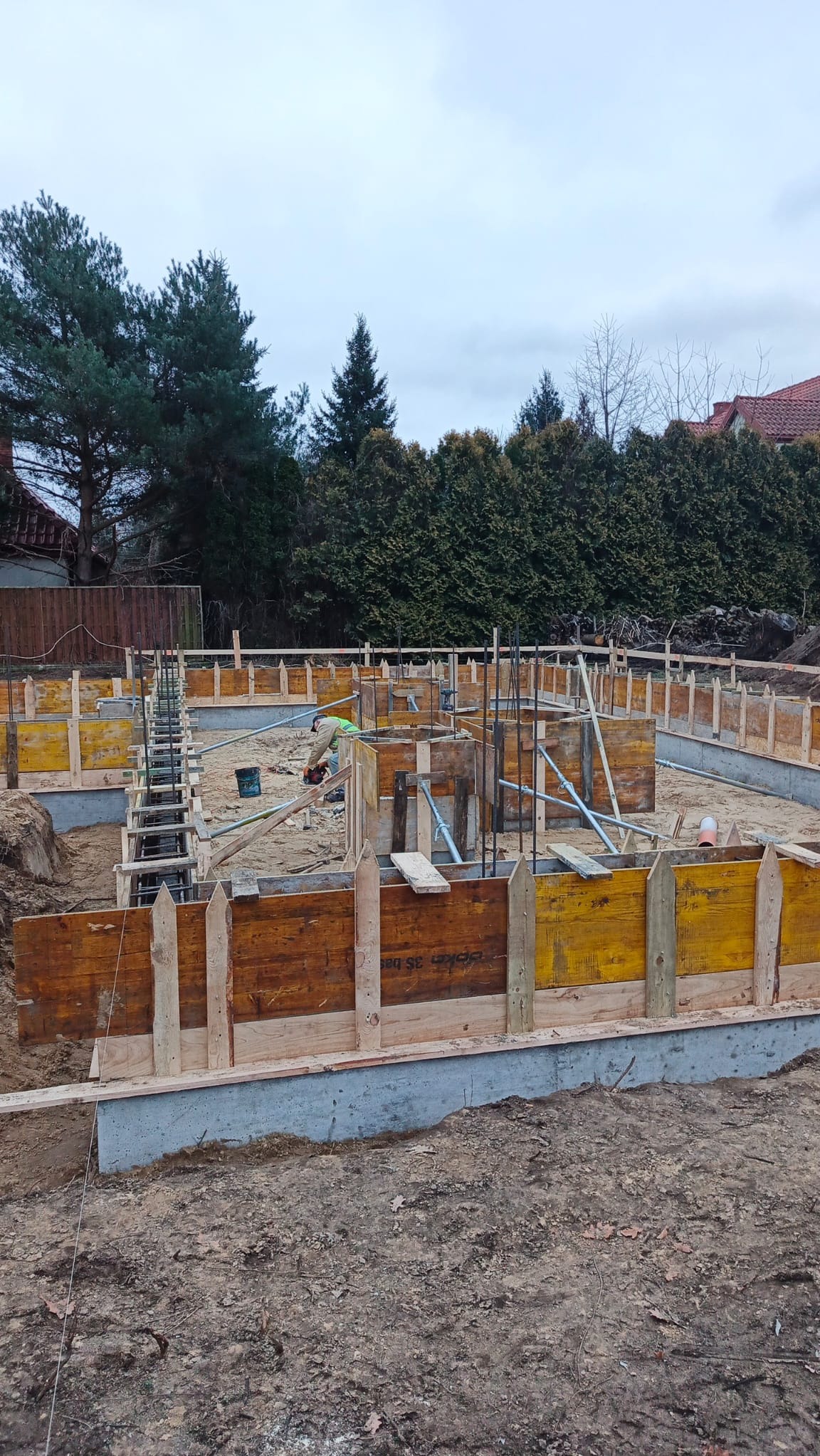 Wykopane fundamenty z drewnianym szalunkiem i zbrojeniem, przygotowane do zalania betonem. W tle zieleń drzew i zabudowa jednorodzinna. Prace ziemne w toku.