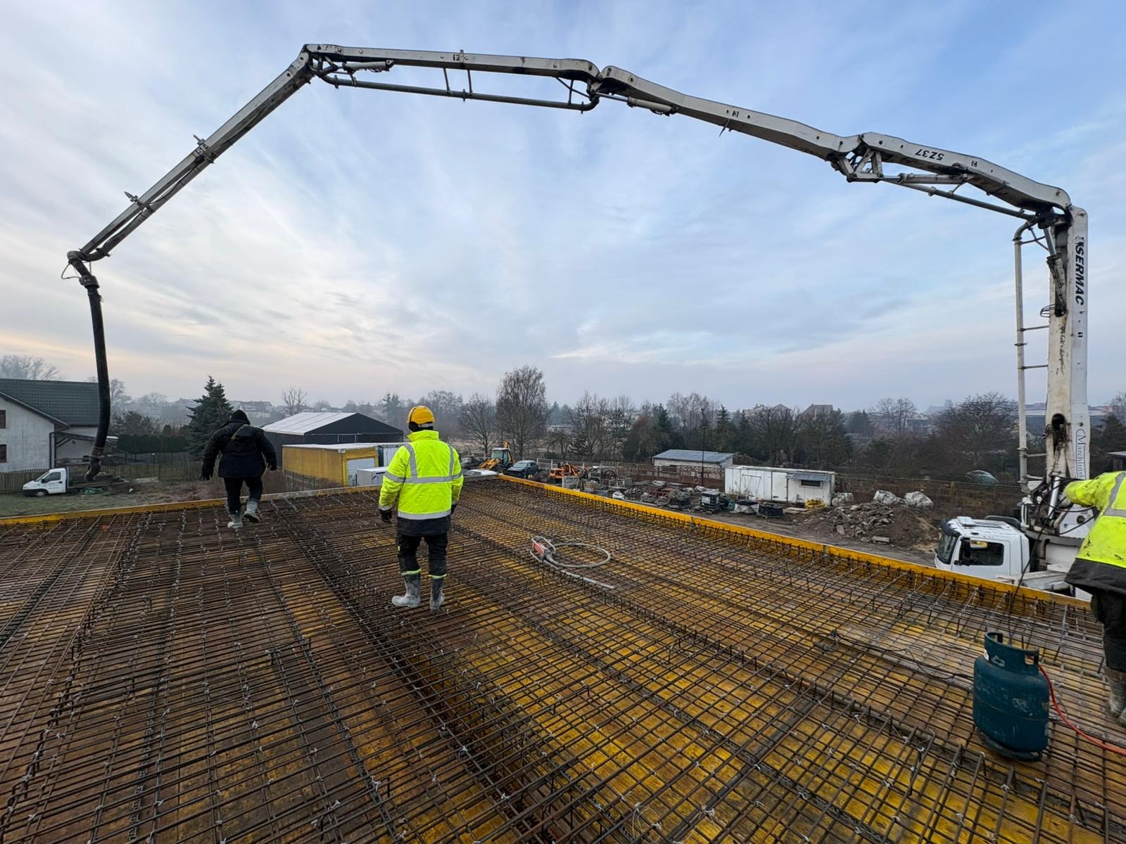 Zbrojenie stropu budynku przygotowane do zalewania betonem. Widoczna pompa do betonu i pracownicy w odzieży roboczej. Żółte szalunki i stalowe pręty tworzą siatkę.