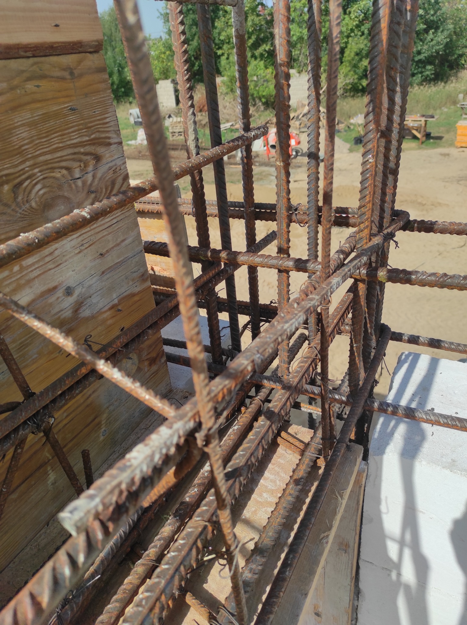 Zbrojenie pionowe z żebrowanych prętów stalowych, połączonych drutem, tworzących szkielet konstrukcji, widoczne drewniane szalunki i fragment białego bloczka, w tle plac budowy z betoniarką.