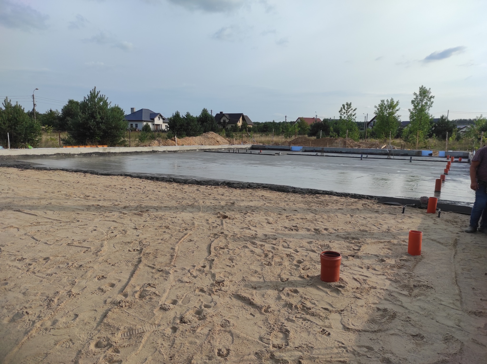 Wylany fundament pod budowę domu jednorodzinnego, widoczne szalunki, zbrojenie i wystające rury kanalizacyjne, w tle zabudowa mieszkalna.