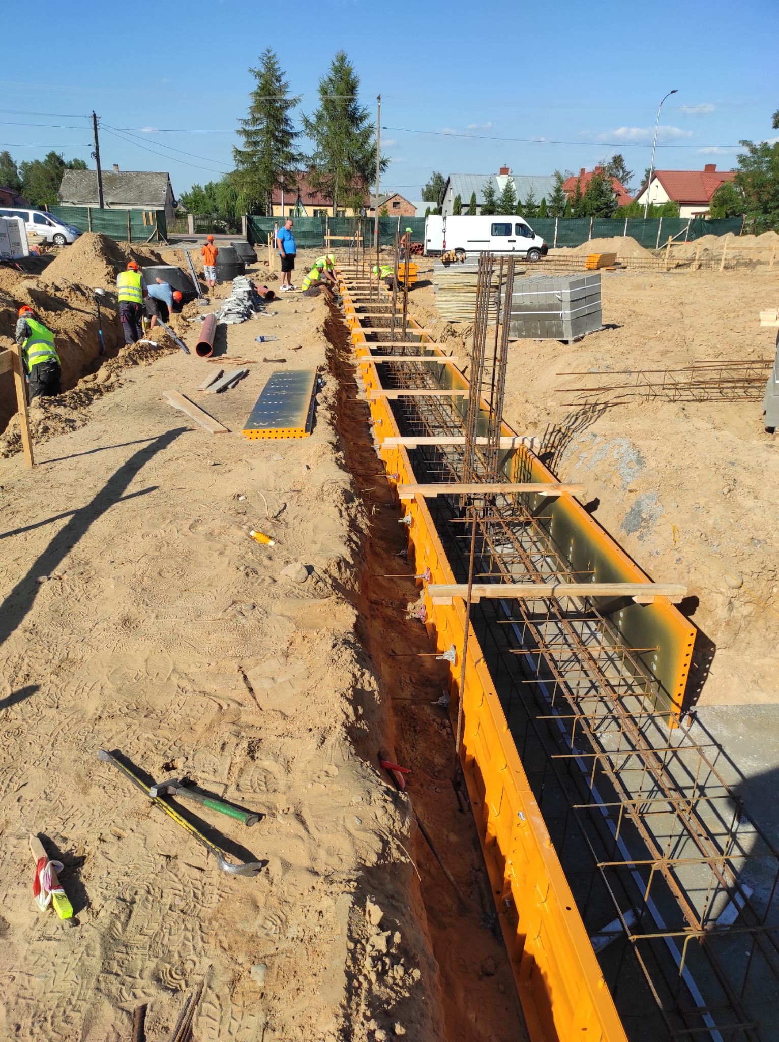 Przygotowanie szalunków i zbrojenia do wylewania fundamentów na placu budowy, widoczni pracownicy w odblaskowych kamizelkach, w tle domy jednorodzinne.