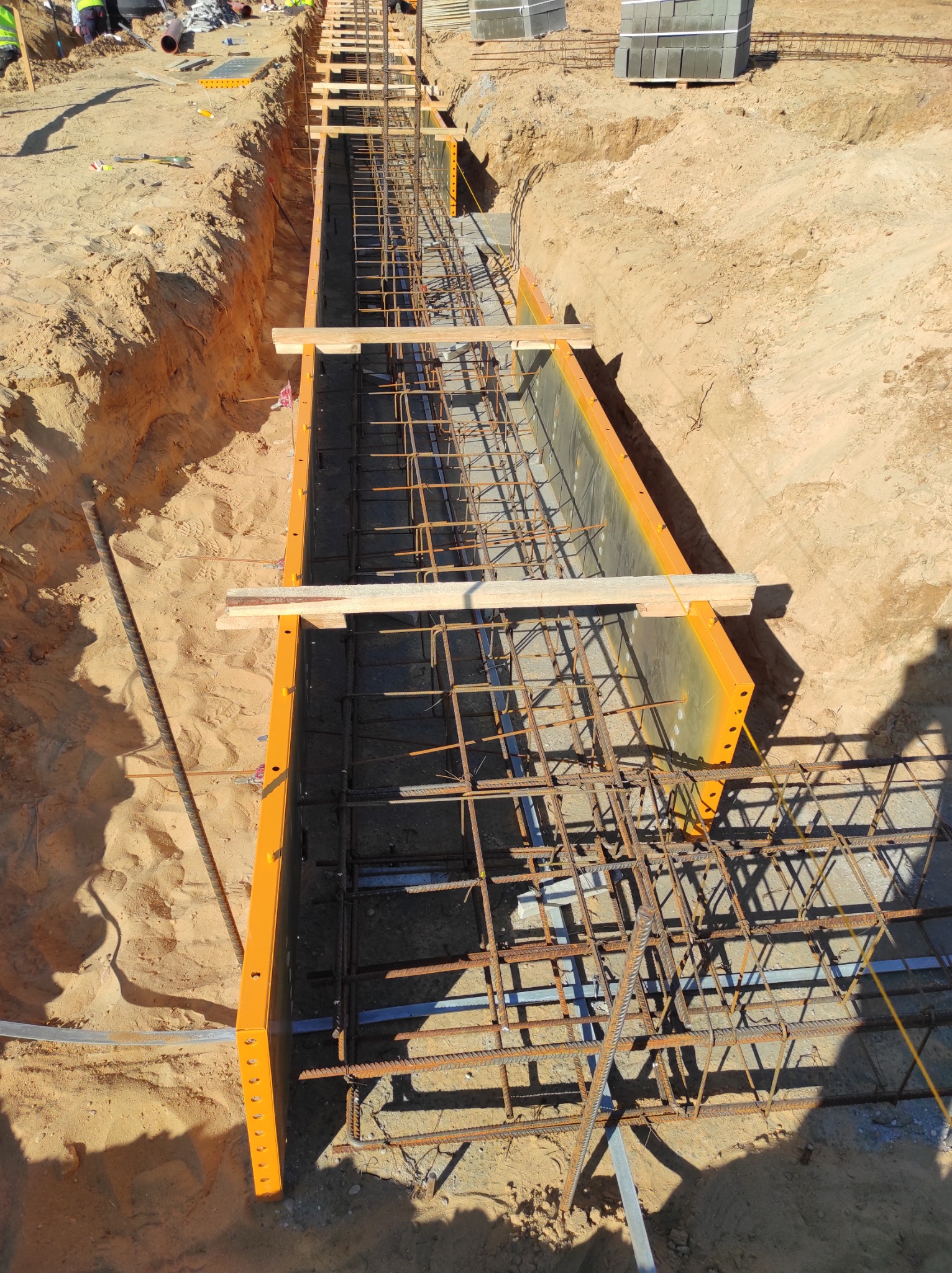 Przygotowanie szalunku i zbrojenia pod wylewkę fundamentów, widoczne drewniane rozpórki i elementy stalowe, ziemia wokół wykopu.