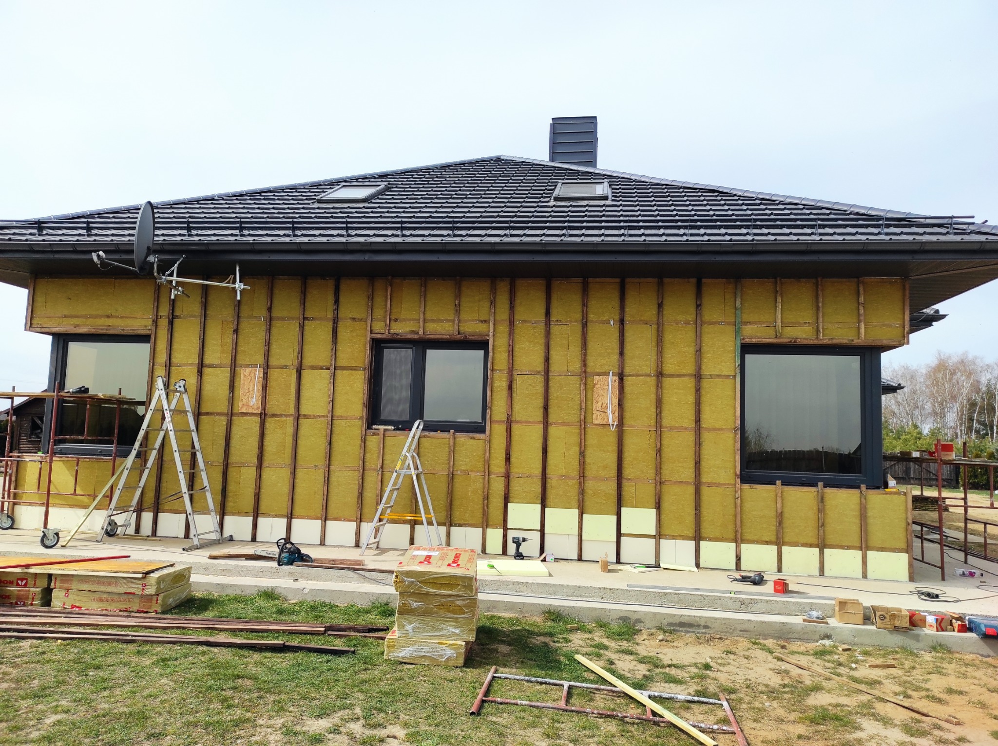 Dom w trakcie ocieplania elewacji, widoczne rusztowania, drabiny, paczki wełny mineralnej, częściowo zamontowane płyty izolacyjne, czarne ramy okienne i satelita na dachu.