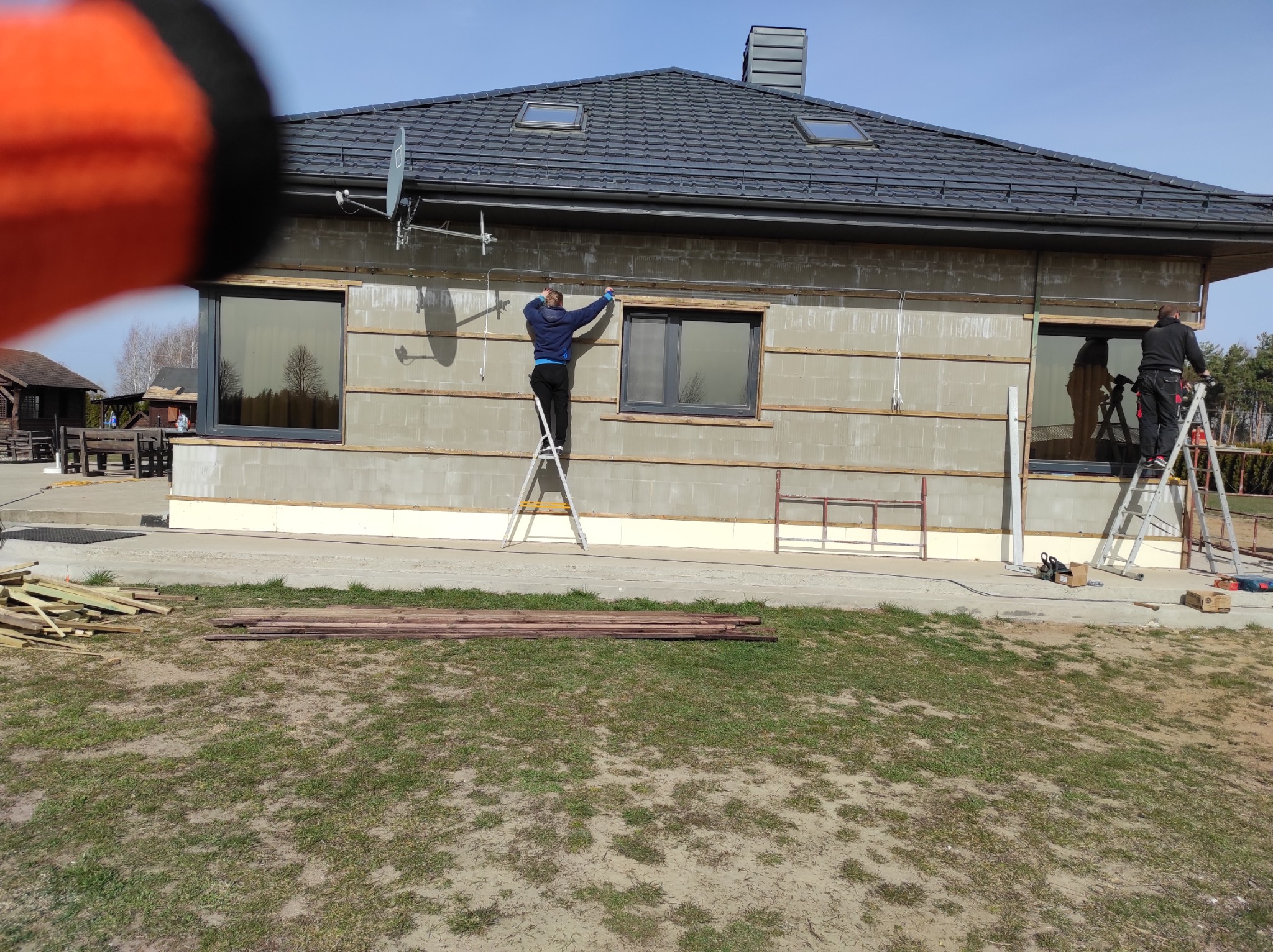 Dwóch pracowników na drabinach montuje listwy na ścianie zewnętrznej domu w trakcie budowy; widoczne okna, dach z dachówkami i elementy instalacji satelitarnej.