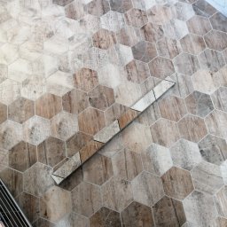 ProTejgels Adrian Dąbrowski - Podłoga wyłożona heksagonalnymi płytkami imitującymi drewno w odcieniach brązu i szarości, z widoczną listwą wykończeniową i fragmentem metalowego stojaka w lewym dolnym rogu.