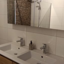 ProTejgels Adrian Dąbrowski - Nowoczesna łazienka z podwójną umywalką nablatową na drewnianej szafce, białe płytki na ścianach i lustro z widoczną kabiną prysznicową w odbiciu.