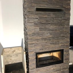 ProTejgels Adrian Dąbrowski - Obudowa kominka wykończona kamieniem dekoracyjnym w odcieniach szarości i brązu, obok drewniany pojemnik na drewno na kółkach, czarna podłoga.
