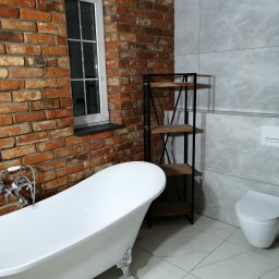 ProTejgels Adrian Dąbrowski - Nowoczesna łazienka z wolnostojącą wanną na lwich nóżkach, ceglaną ścianą, szarą płytką imitującą marmur, białą ceramiką i metalowym regałem z drewnianymi półkami.