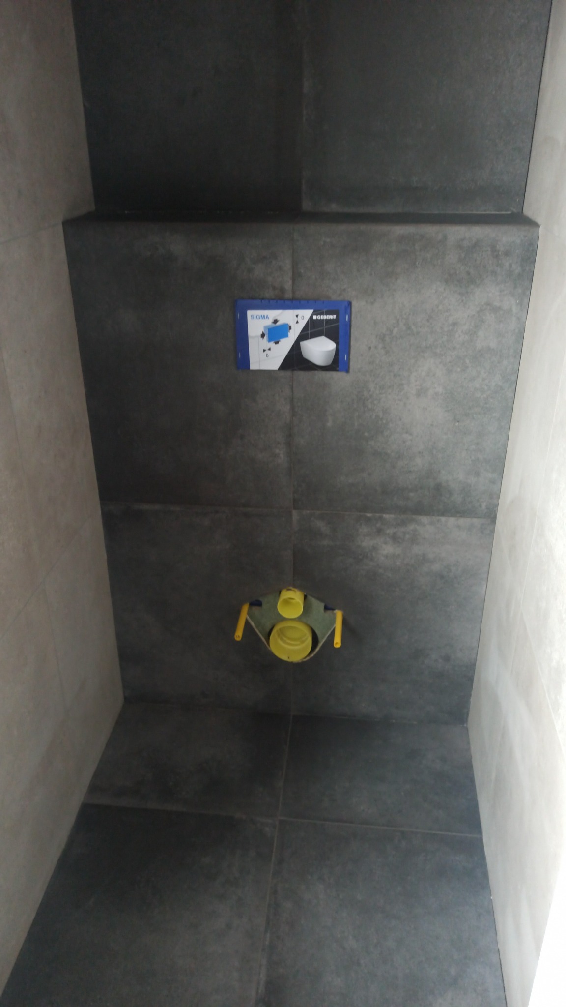 Szara ściana w łazience w Zbąszyniu wyłożona płytkami, z widoczną instalacją podtynkową WC (Geberit Sigma) przed montażem miski ustępowej.