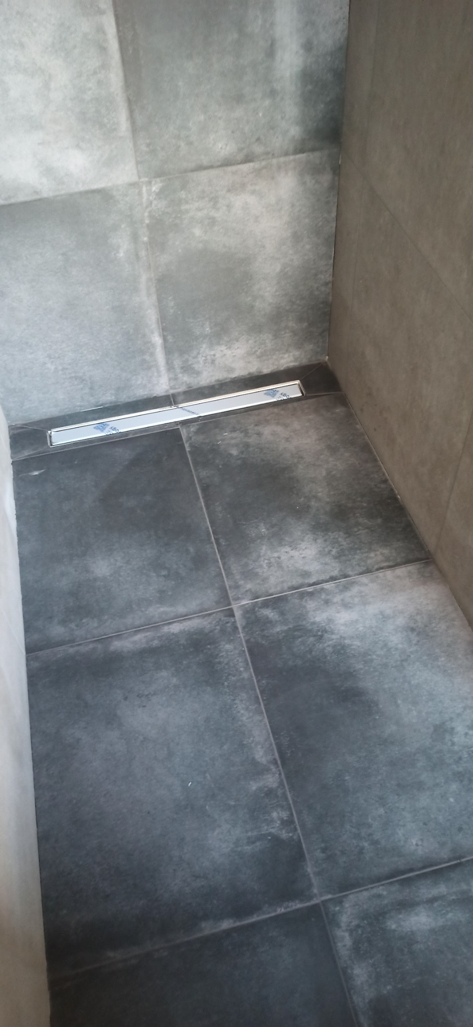 Szare płytki podłogowe i ścienne w kabinie prysznicowej z liniowym odpływem podłogowym, zabezpieczonym jeszcze folią ochronną.