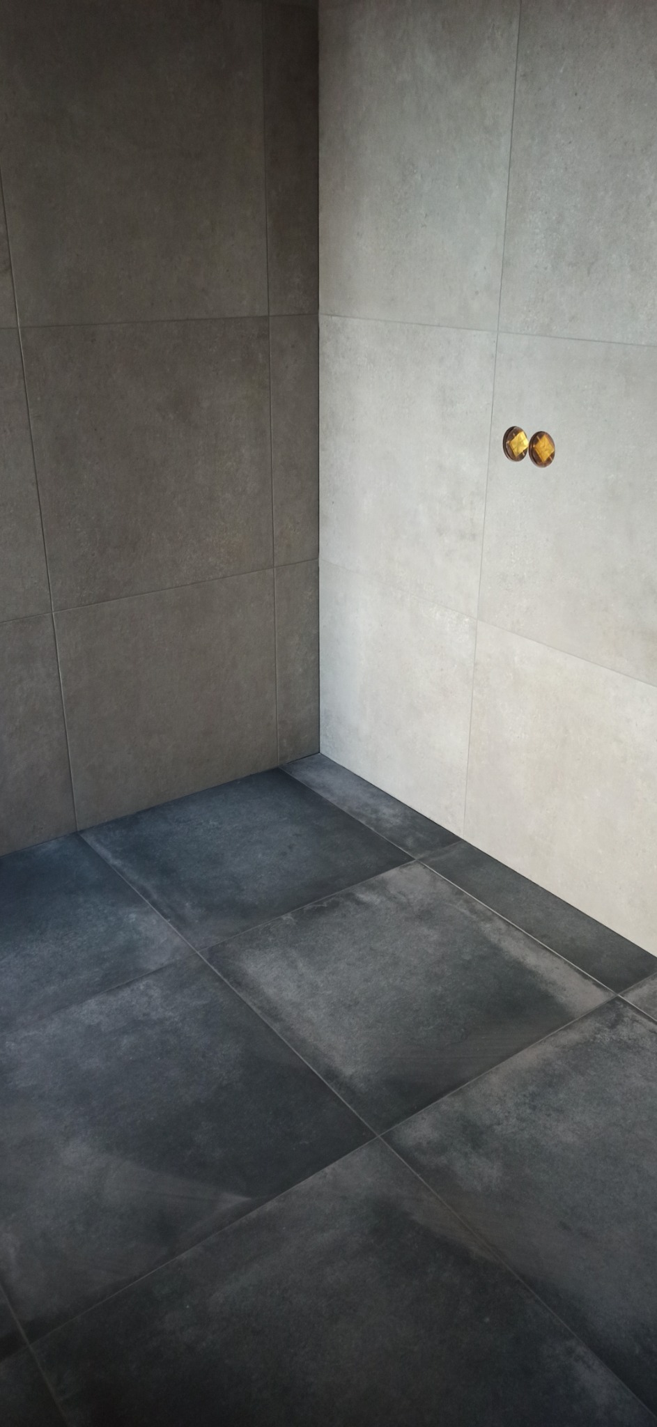 Wykończona łazienka z ciemnymi płytkami podłogowymi i jasnoszarymi płytkami ściennymi z widocznymi przyłączami wodnymi.