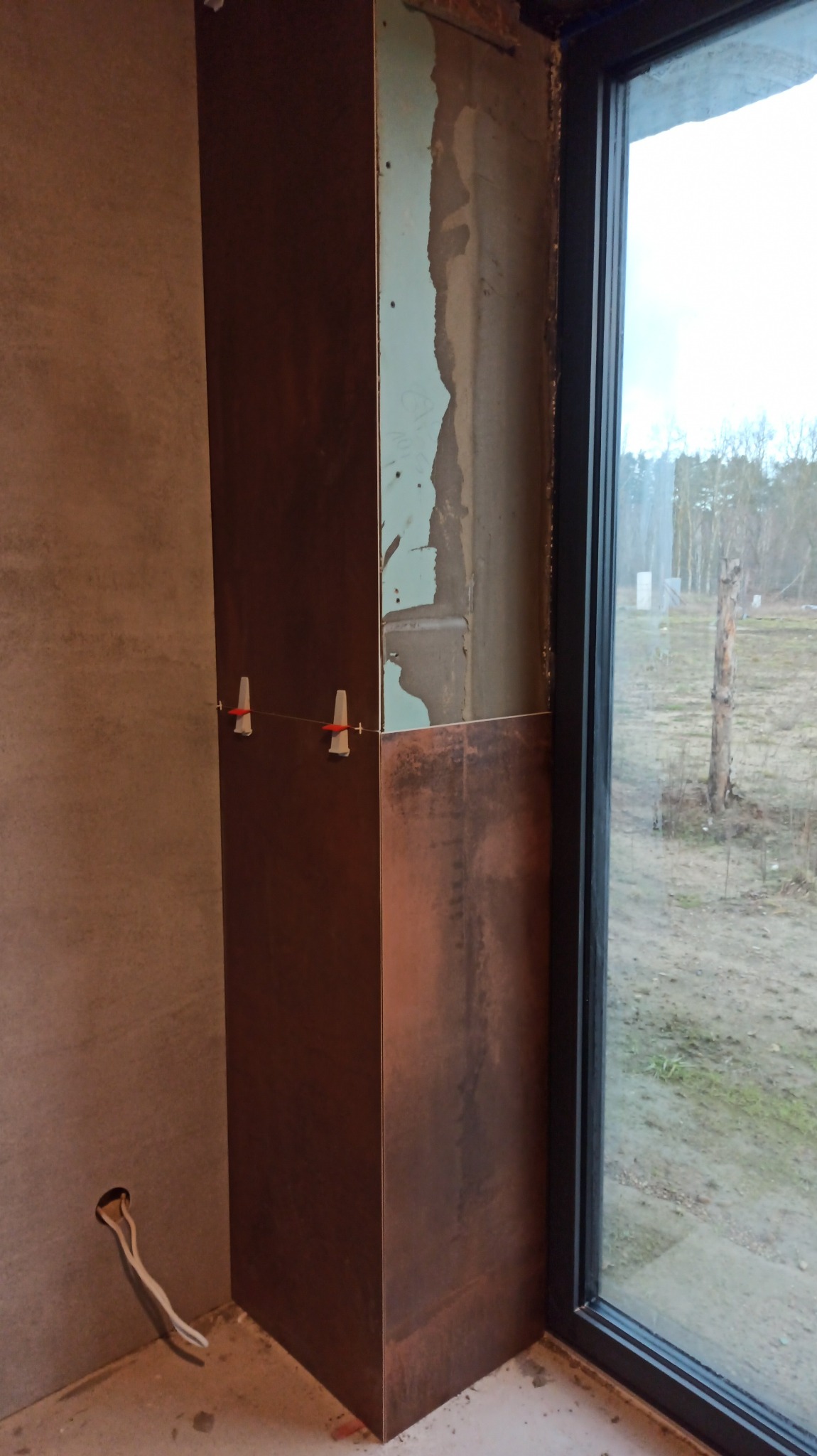 Układanie brązowych płytek imitujących rdzę na narożniku ściany przy oknie, z widocznymi krzyżykami dystansowymi i fragmentem nieotynkowanej ściany obok płyty kartonowo-gipsowej.