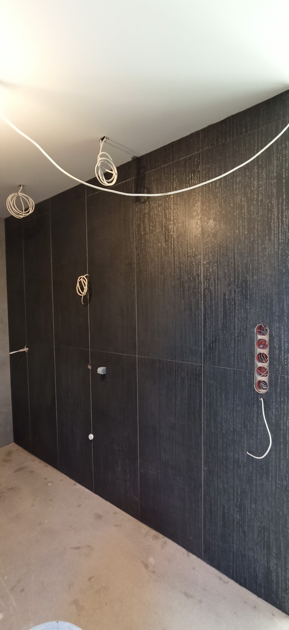 Ściana wyłożona ciemnymi płytkami z widocznymi kablami elektrycznymi, gniazdkami i instalacją hydrauliczną, w trakcie prac wykończeniowych.