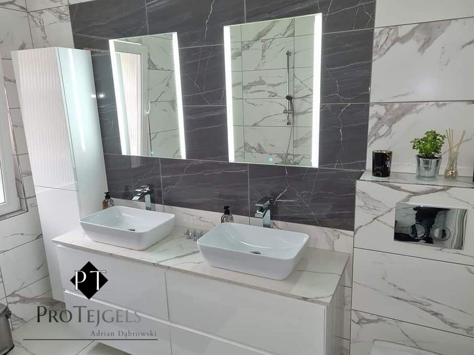 Nowoczesna łazienka z dwoma białymi umywalkami nablatowymi, podświetlanymi lustrami i marmurowymi płytkami w odcieniach szarości i bieli. Na blacie między umywalkami stoi dozownik mydła. Widoczne...