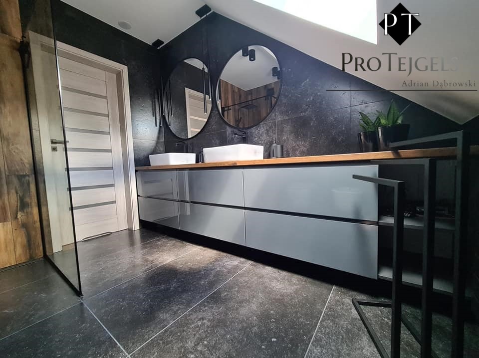 Nowoczesna łazienka na poddaszu z szarą szafką pod umywalkami, drewnianym blatem, okrągłymi lustrami, czarnymi płytkami na ścianie i podłodze oraz przeszkloną kabiną prysznicową.