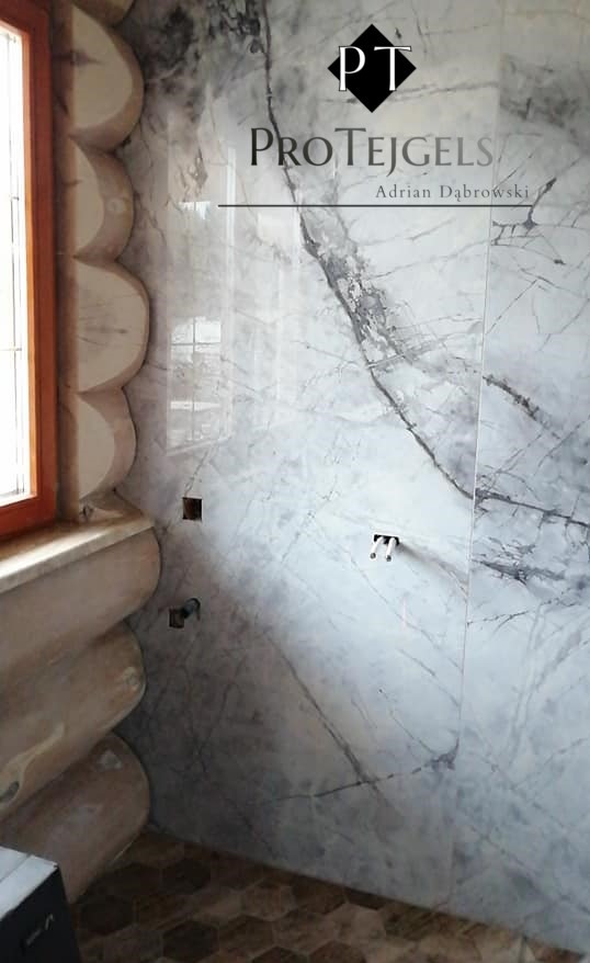 Wykończona łazienka w stylu rustykalnym z marmurowymi płytkami na ścianach, drewnianym oknem i heksagonalną podłogą, widoczne logo firmy na ścianie.