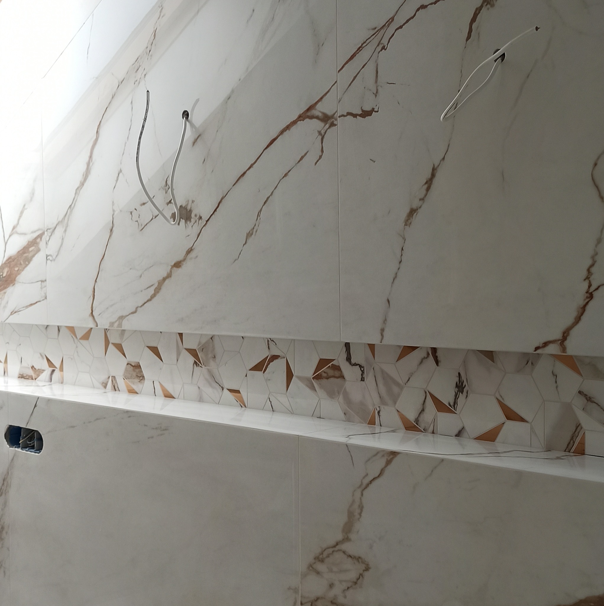 Ściana wyłożona białymi płytkami imitującymi marmur z brązowymi żyłami, z dekoracyjnym pasem heksagonalnych płytek z brązowymi akcentami, widoczne wystające kable elektryczne.