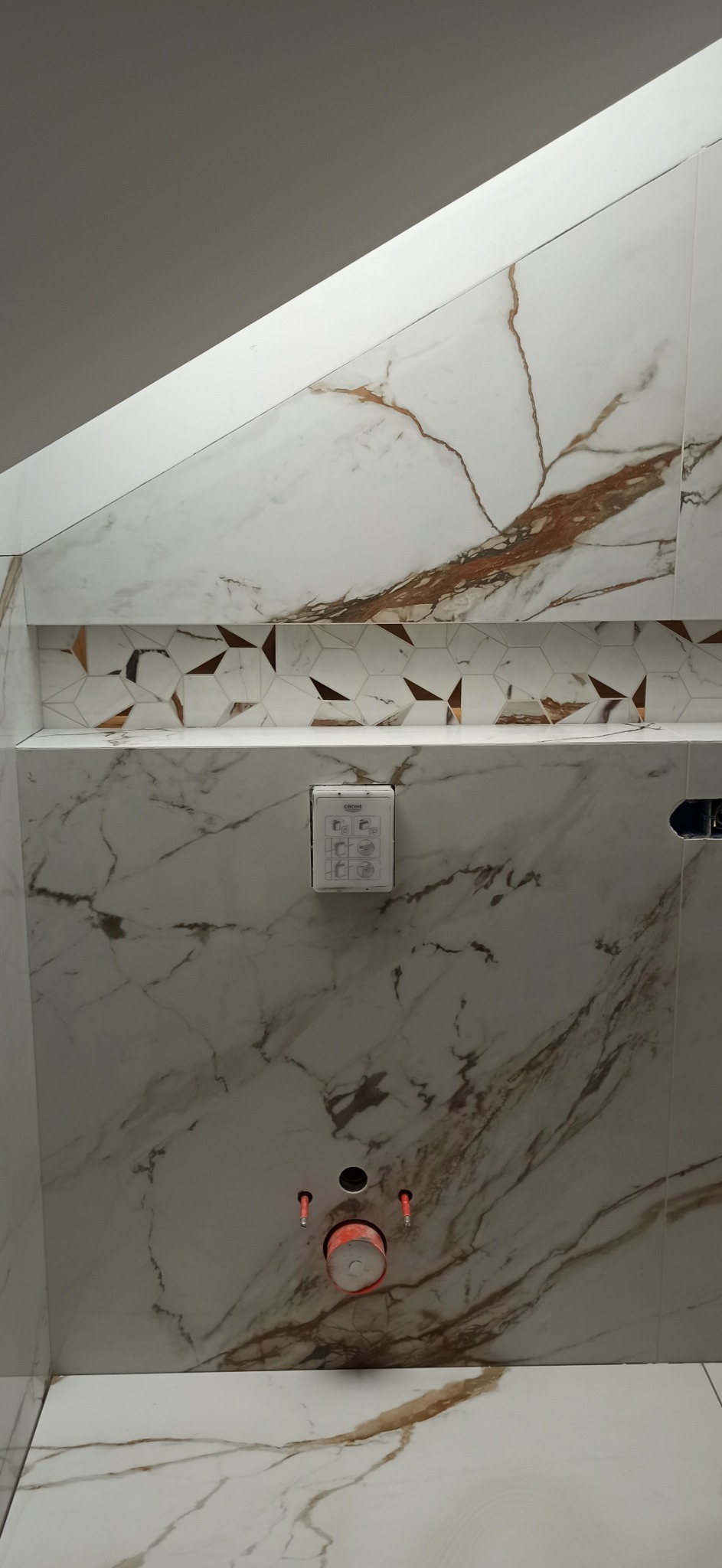 Ściana łazienki wyłożona białymi płytkami imitującymi marmur z brązowymi żyłkami. Nad zabudową stelaża WC dekoracyjny pas z płytek heksagonalnych. Widoczne przyłącza wodne.