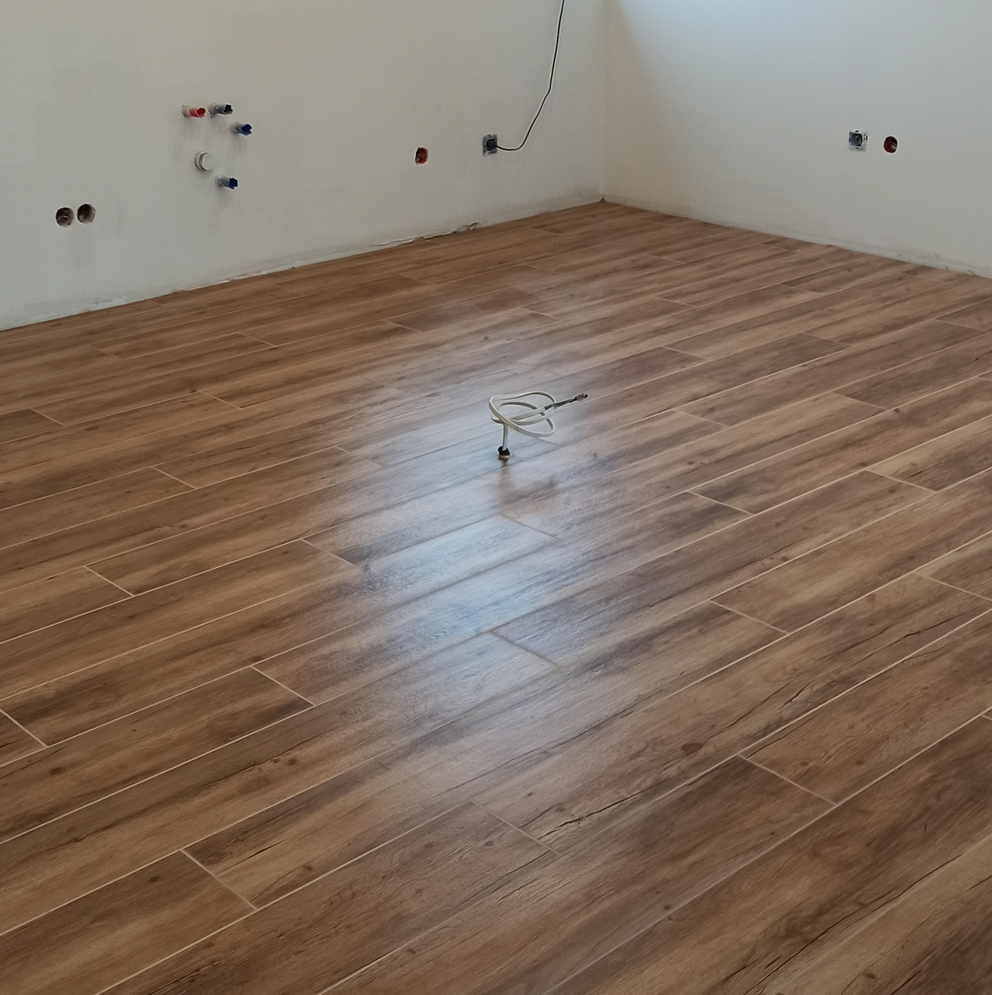 Ułożona podłoga z płytek imitujących drewno w pustym pomieszczeniu z wystającymi instalacjami wodnymi i elektrycznymi w ścianie.