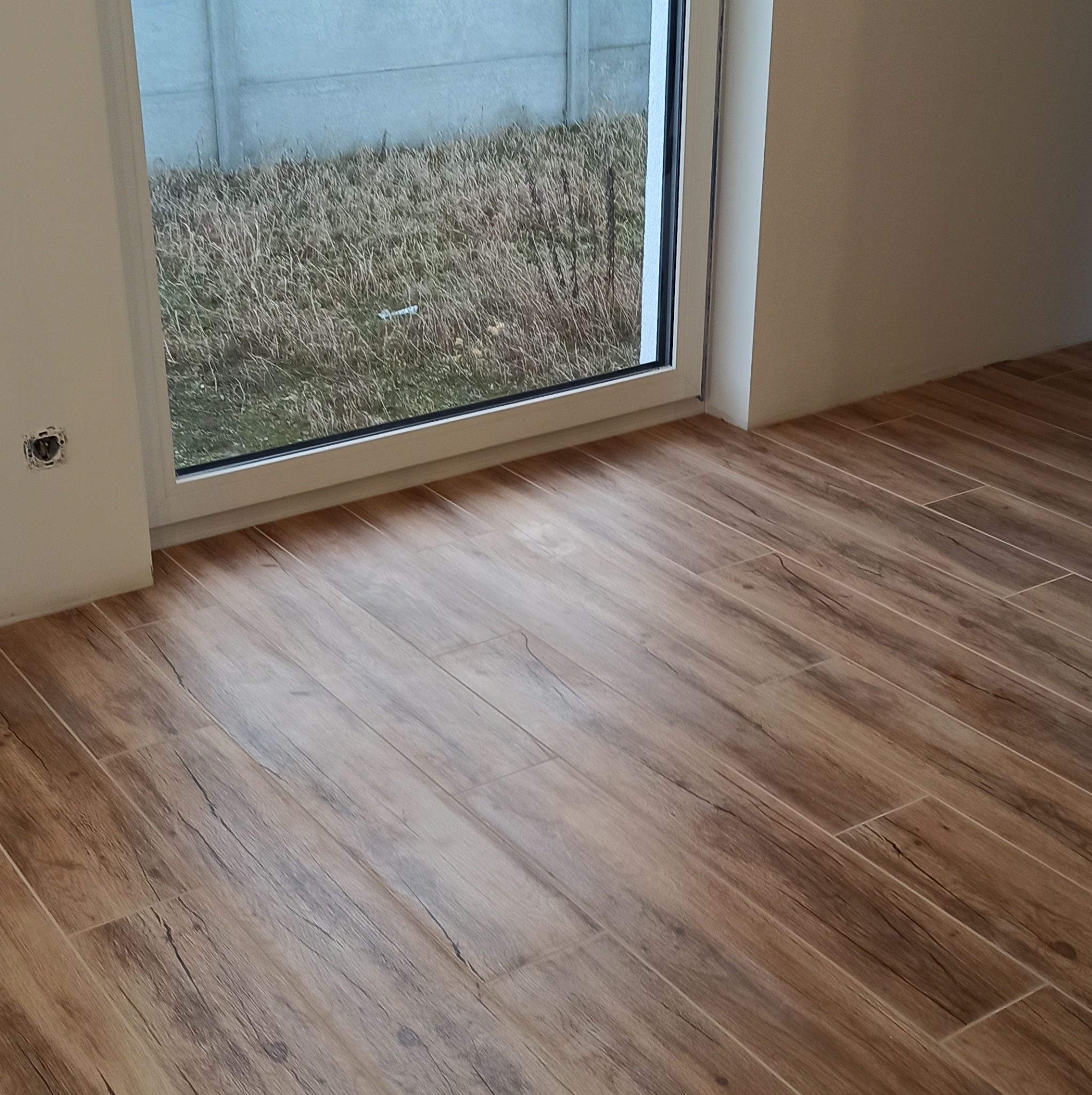 Ułożona pod kątem brązowa podłoga z płytek imitujących drewno, widok na okno z widokiem na trawę i betonowy płot.