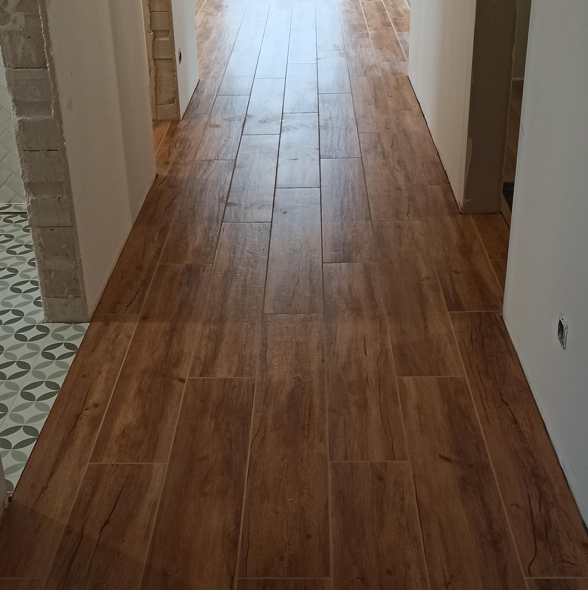 Ułożona podłoga z płytek imitujących drewno w korytarzu, widoczne surowe wykończenia ścian i fragment dekoracyjnej podłogi w sąsiednim pomieszczeniu.