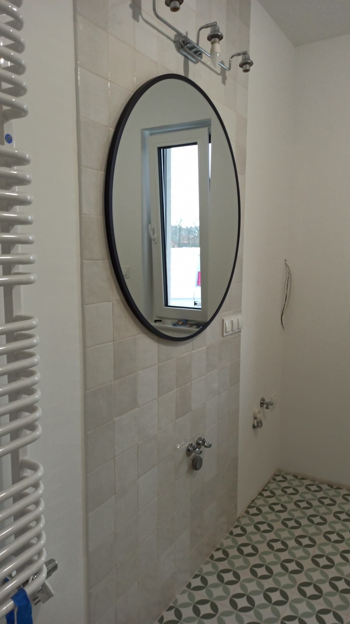 Jasna łazienka z owalnym lustrem, grzejnikiem drabinkowym, wykończona kwadratowymi płytkami i wzorzystą podłogą. Widoczne elementy instalacji wodnej.