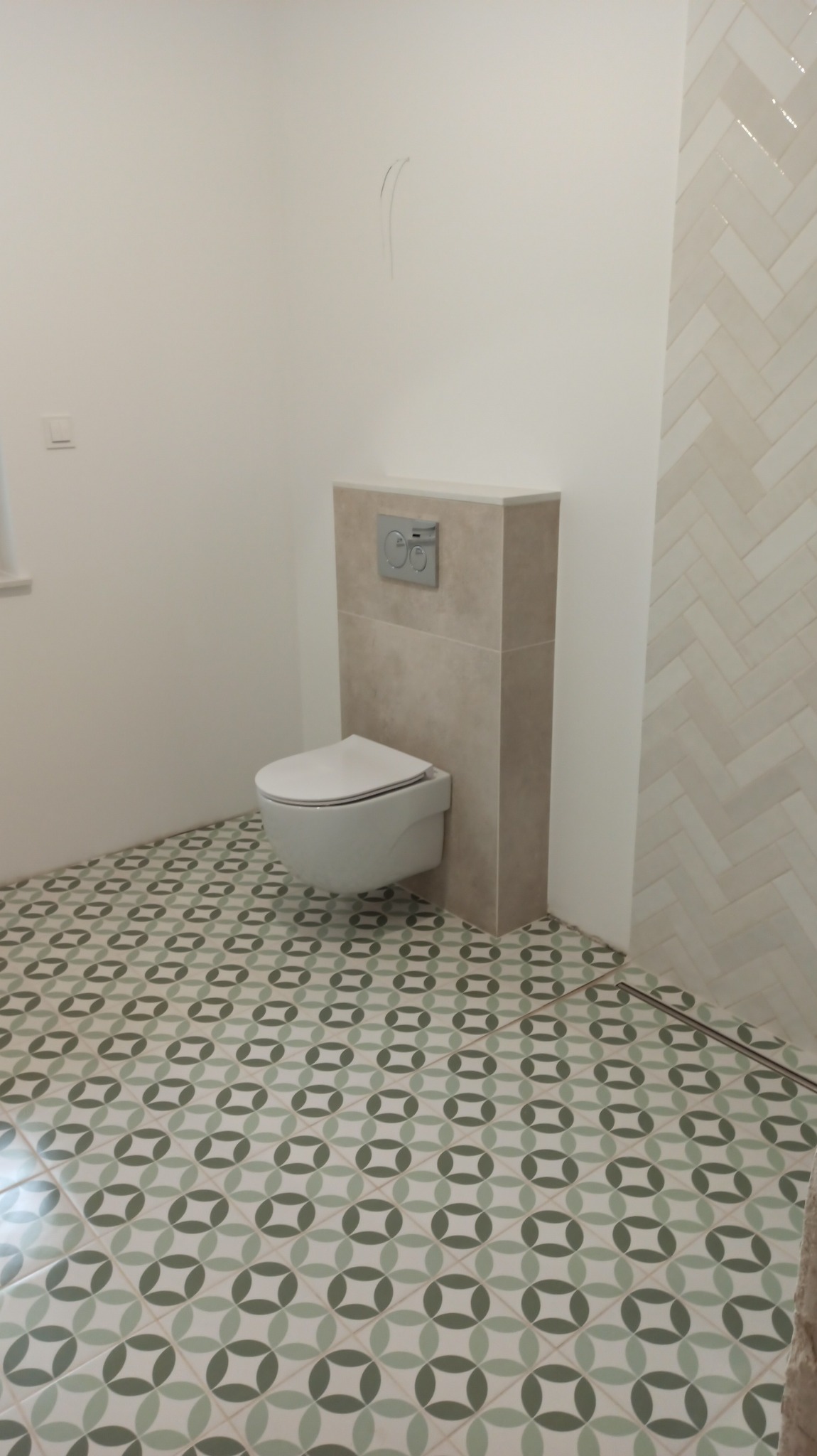 Nowoczesna toaleta podwieszana z zabudową geberit w beżowej okładzinie, wzorzysta podłoga w geometryczne motywy oraz ściana wyłożona białymi płytkami w jodełkę.