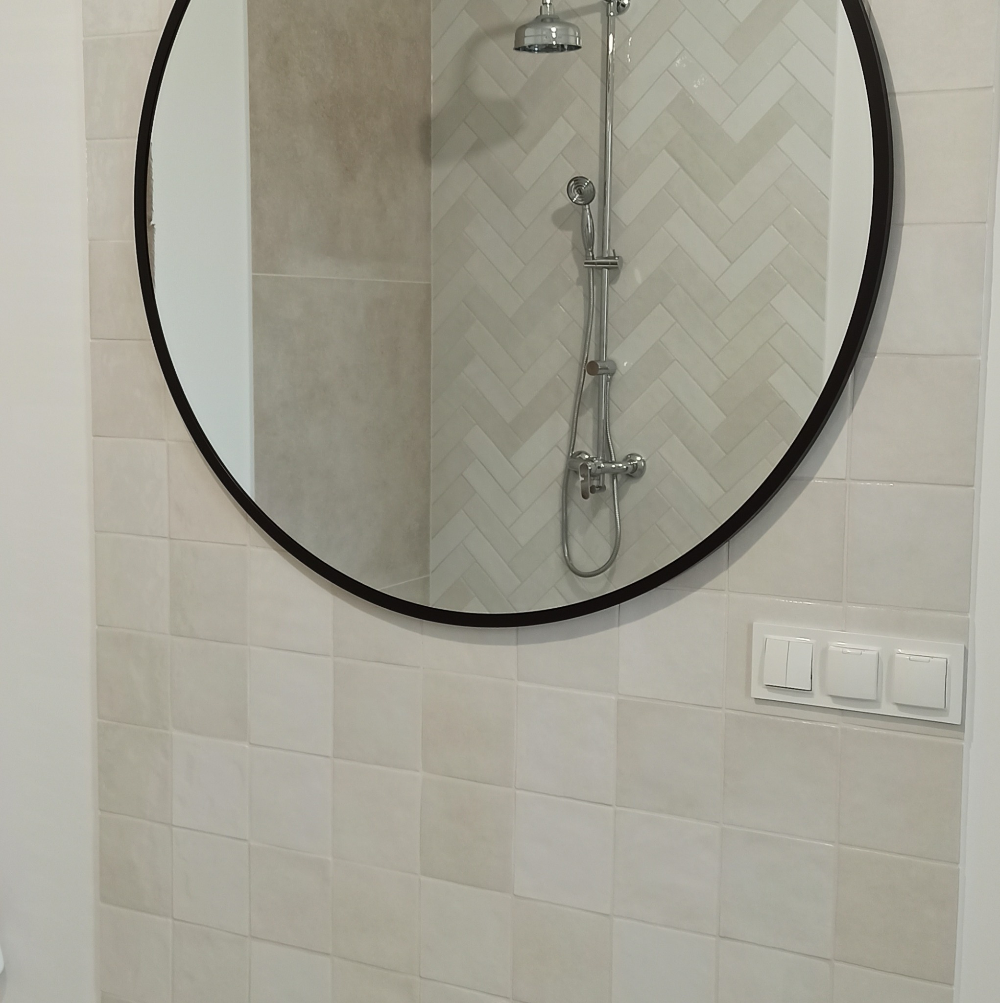 Okrągłe lustro z czarną ramą odbija fragment kabiny prysznicowej z szarymi płytkami ułożonymi w jodełkę, widoczne białe płytki kwadratowe na ścianie i panel z trzema włącznikami światła.