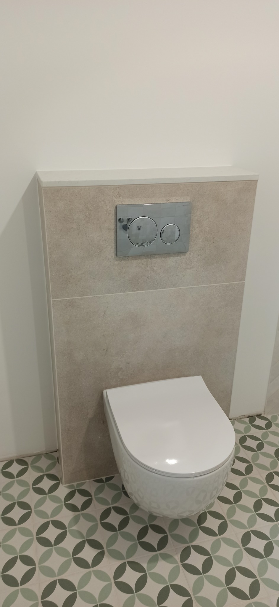 Wisząca toaleta z chromowanym panelem spłukującym na tle beżowych płytek i wzorzystej podłogi w odcieniach zieleni.