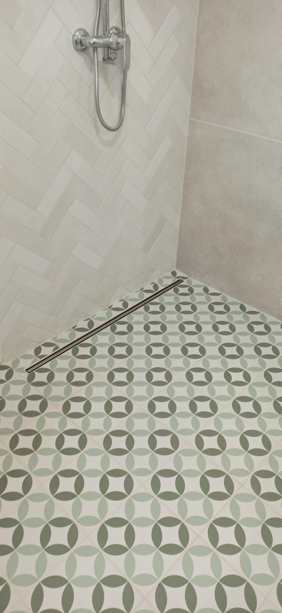 Wykończona kabina prysznicowa z odpływem liniowym, ściany wyłożone białymi płytkami ułożonymi w jodełkę i płytkami w odcieniach beżu, podłoga z wzorem geometrycznym w kolorach zieleni i bieli.