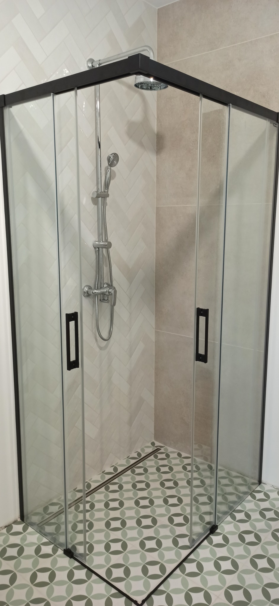 Kabina prysznicowa z czarnymi ramami i deszczownicą, ściany wyłożone płytkami w dwóch odcieniach beżu, podłoga z wzorem geometrycznym w odcieniach zieleni i szarości.