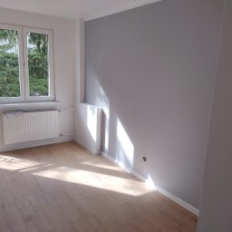 Lech-Bud - Pomalowane na szaro wnętrze pokoju z parkietem, grzejnikiem i oknem z widokiem na drzewa, oświetlone promieniami słońca