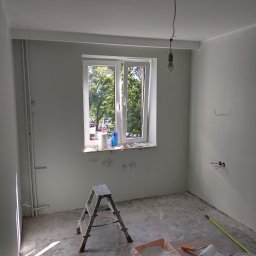 Lech-Bud - Pokój w trakcie prac malarskich, widoczna drabina, puszki z farbą, wałki i kuwety malarskie, ściany i sufit świeżo pomalowane na biało, okno z widokiem na zieleń na zewnątrz, surowa żarówka...