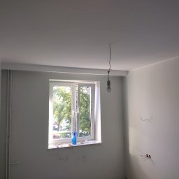 Lech-Bud - Wnętrze pokoju po malowaniu na biało, widoczne świeżo pomalowane ściany i sufit, okno z widokiem na zieleń, na parapecie niebieski spryskiwacz i resztki taśmy malarskiej, wystające kable elektryczne.