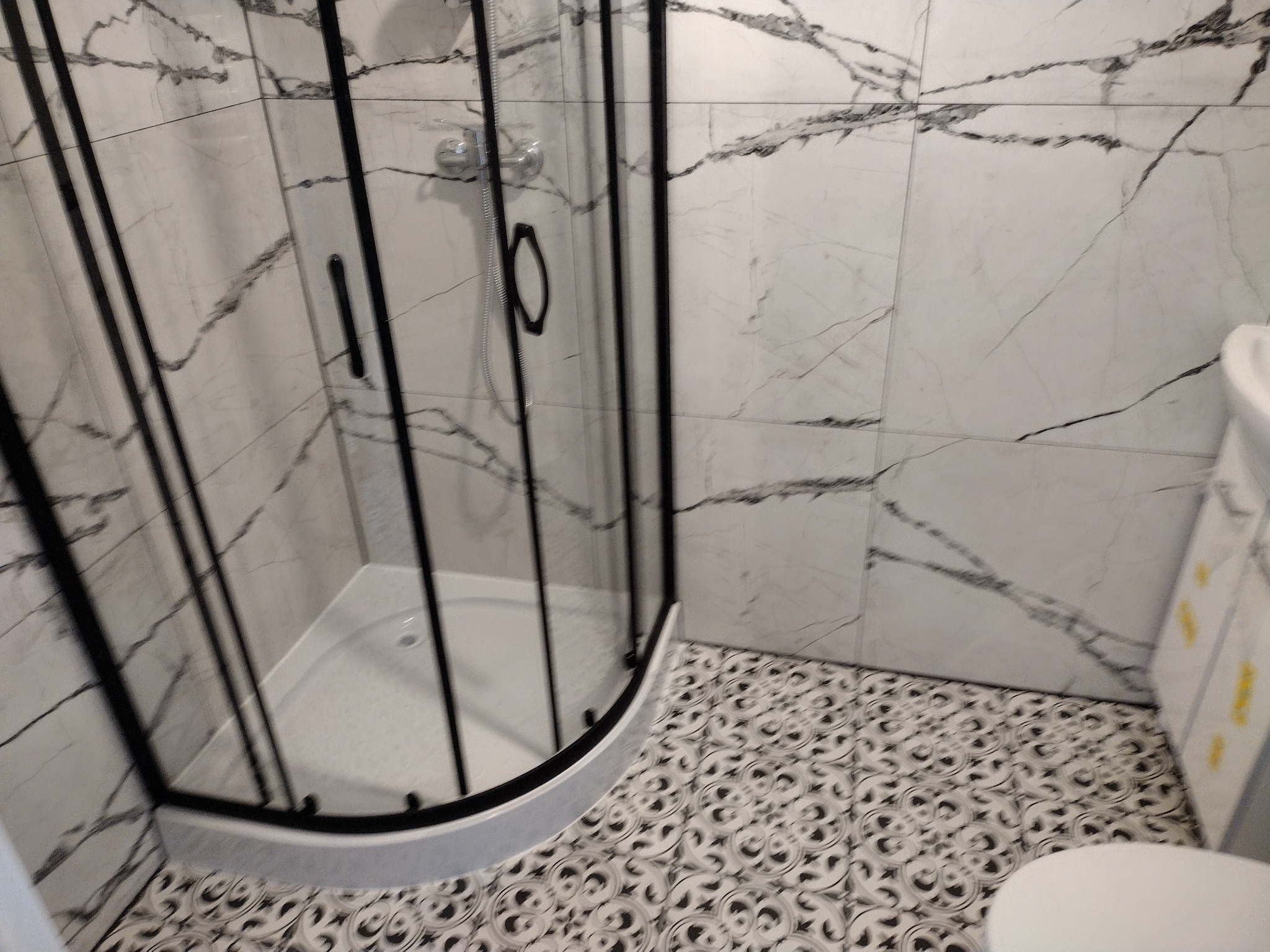 Narożna kabina prysznicowa z czarnymi profilami na tle białych płytek imitujących marmur i wzorzystej podłogi.