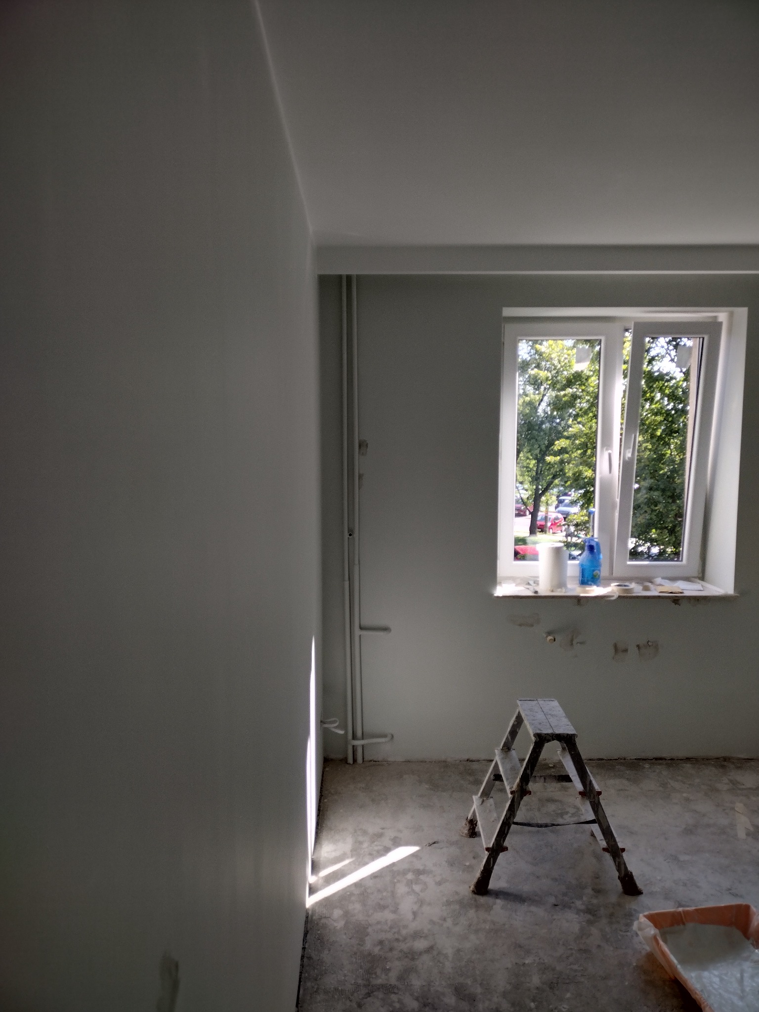 Pokój w trakcie odświeżania: ściana po nałożeniu gładzi, drabina malarska, kuweta z farbą, okno z widokiem na zieleń, ślady po szpachlowaniu pod oknem.