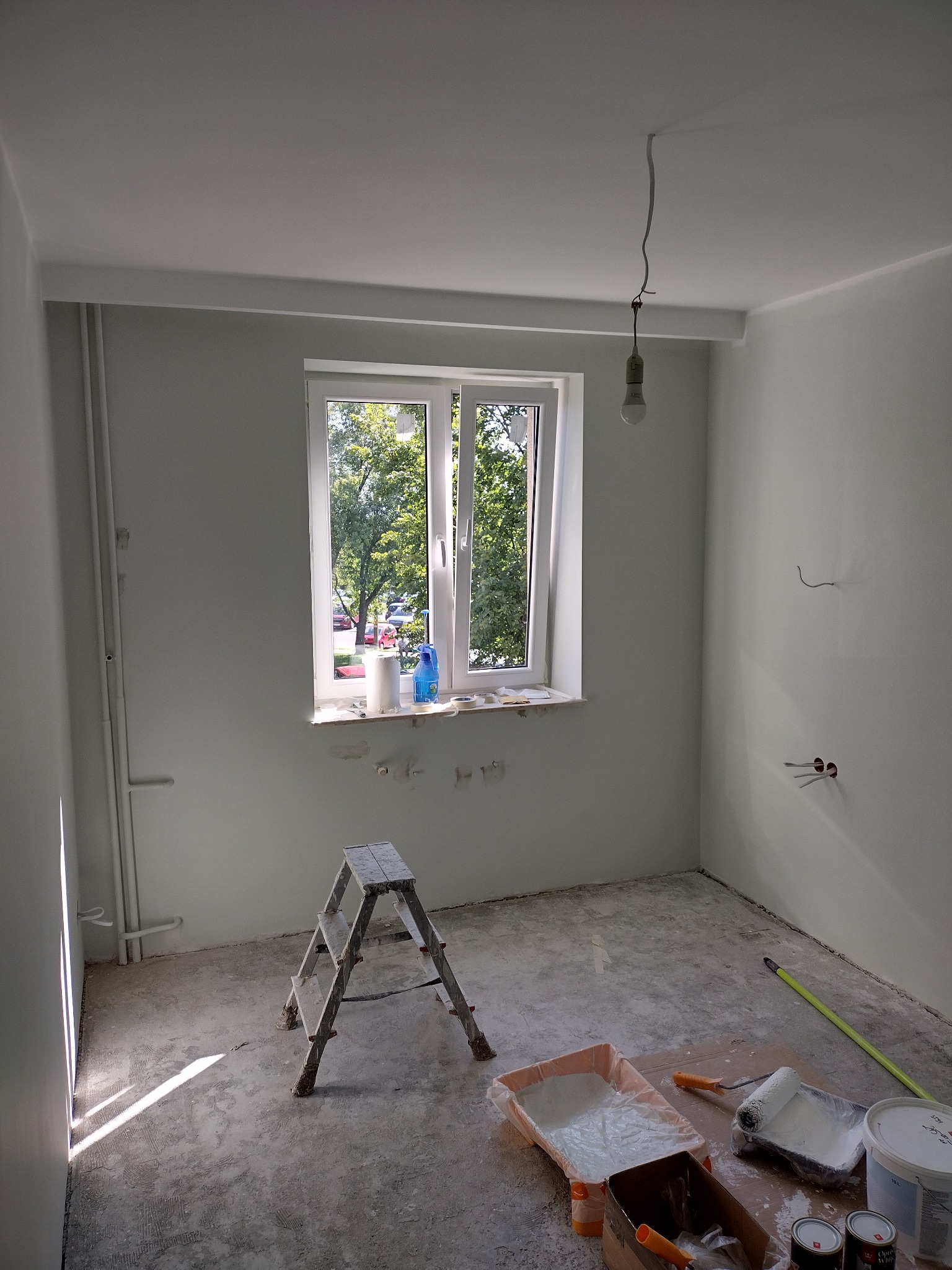 Pokój w trakcie prac malarskich, widoczna drabina, puszki z farbą, wałki i kuwety malarskie, ściany i sufit świeżo pomalowane na biało, okno z widokiem na zieleń na zewnątrz, surowa żarówka...