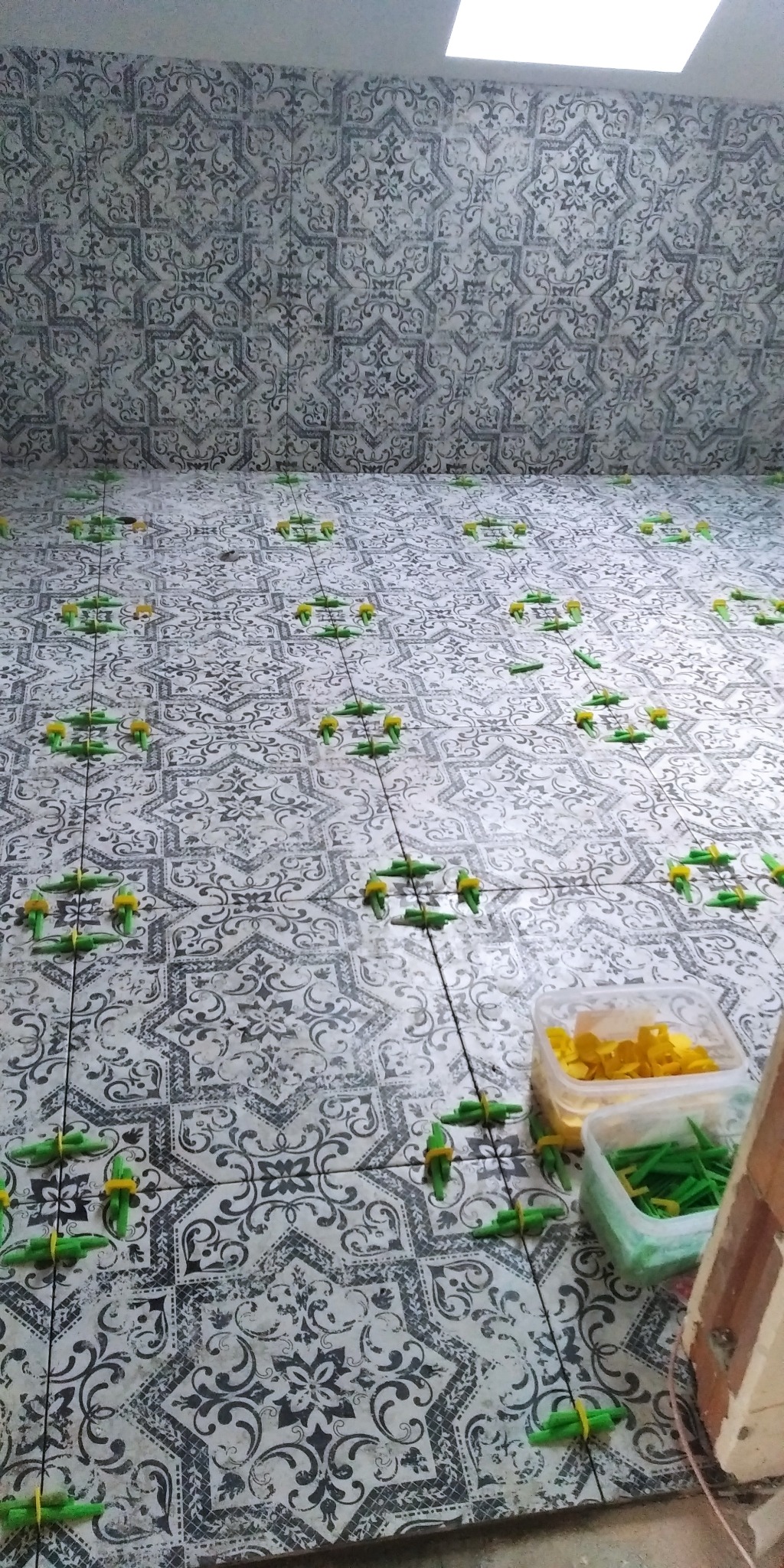 Układanie wzorzystych płytek podłogowych z użyciem systemów poziomowania, widoczne pojemniki z klinami i klipsami.