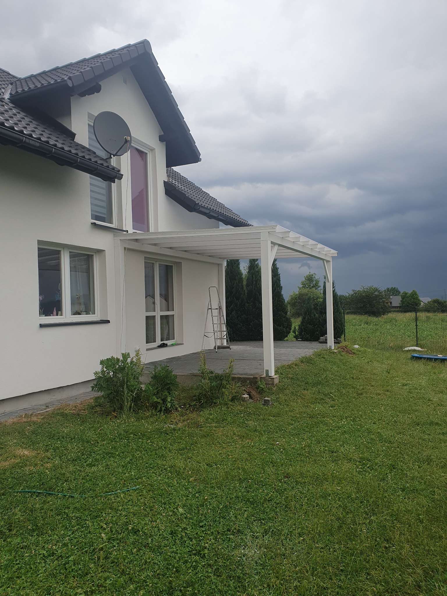 Biała, drewniana konstrukcja przylegająca do ściany domu z anteną satelitarną, na tle burzowego nieba i zielonego trawnika. Widoczna drabina oparta o ścianę.