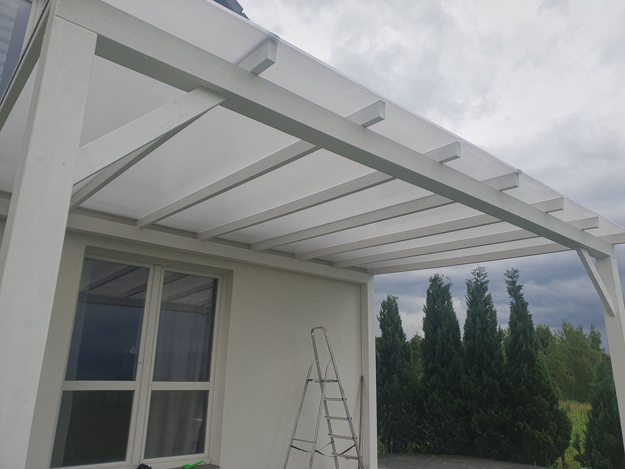 Biała altana z przezroczystym zadaszeniem przylegająca do budynku, drabina oparta o ścianę, widok na iglaki i zachmurzone niebo.