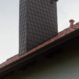 Nowoczesny komin obłożony łupkiem w kolorze grafitowym, z kratkami wentylacyjnymi u góry, wystający ponad dach z czerwoną dachówką ceramiczną, na tle pochmurnego nieba.