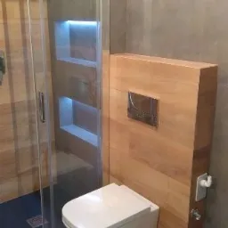 Nowoczesna łazienka z podwieszaną toaletą na drewnianej zabudowie, szklaną kabiną prysznicową z wnękami podświetlonymi LED, wykończona płytkami imitującymi drewno.