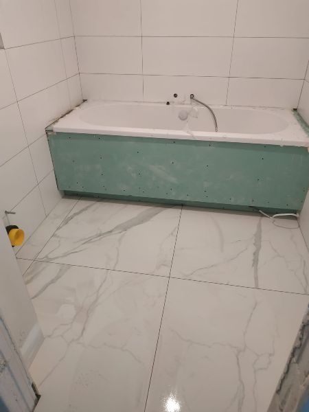 Wanna obudowana płytami gipsowo-kartonowymi, białe płytki na ścianach i podłodze imitujące marmur, widoczne elementy instalacji hydraulicznej przed wykończeniem.