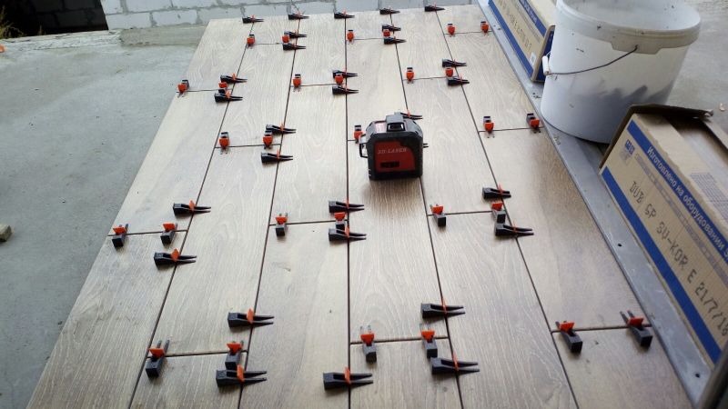 Brązowe płytki podłogowe z systemem poziomowania i laserowym niwelatorem, widoczne pudełka i wiadro w tle.